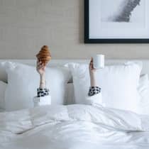 A woman laying under a white plush bedspread holds up both hands, one holding a croissant, another holding a white ceramic mug