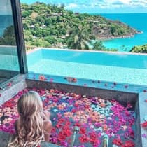 A woman sits in a bathtub full of flower petals by an open window overlooking a private pool, blue waters and a view of the island