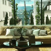 Beige sofa with decorative pillows, a small bonsai plant on a glass coffee table and a glass wall overlooking city buildings, trees and distant mountains