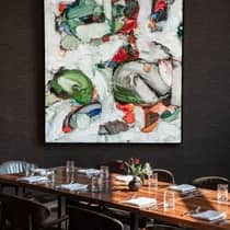 Interior of a dining room with a large wooden table set with white linen napkins, glasses and a floral centerpiece, with a large modern painting on the wall above