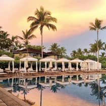 Cabanas, lounge chairs and palm trees line the outdoor pool under a pink and orange sunset. 