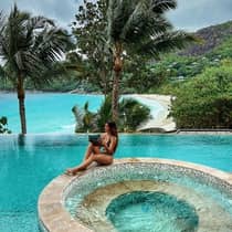 A woman lounges by a beachside pool shaded by palm trees while reading a book