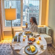 Person in a bathrobe enjoying breakfast in a luxury hotel room with a city view, seated at a round table with an omelette, waffles, fruit and orange juice