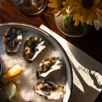 A dish of chilled oysters sits on top of a Goldfinch Tavern menu, next to a glass of white wine and bud vase filled with yellow flowers