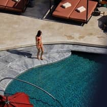 A woman dressed in a red swimsuit walks on marble steps into an outdoor pool surrounded by covered cabanas