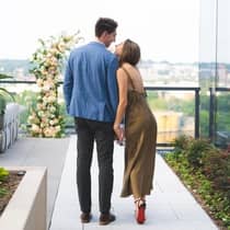 A couple holding hands walking on an outdoor terrace towards a large floral arrangement