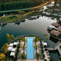 Aerial view of a pool lined with palm trees and cabanas overlooking a lake and trees at sunset