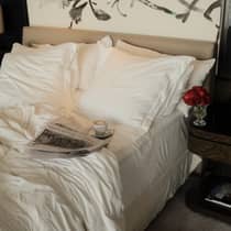 A newspaper and cup of coffee on a saucer sit atop an unmade bed with crisp, white sheets, with a pair of white slippers on the floor beneath it