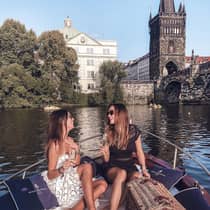 Two people sitting on a boat, enjoying champagne, with a wicker picnic basket and historic buildings and a bridge in the background.