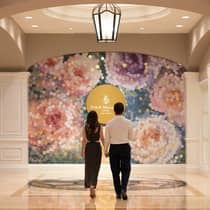A couple dressed in formal attire hold hands while walking towards a colourful mural in an ornate hotel lobby