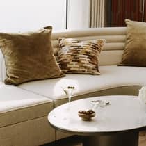 A corner of a room with a tan linen couch with brown accent pillows, a modern, circular coffee table topped with a martini and a bowl of nuts and an abstract printed rug