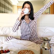 Woman on bed sips coffee cup, stretches in front of in-room breakfast dining tray