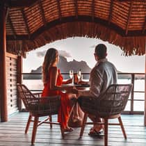 Two people enjoying a sunset view from a waterfront deck with drinks, overlooking a mountain and ocean