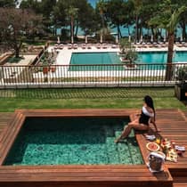 A woman lounges at a plunge pool with various food dishes, beverages and champagne, overlooking a large pool area and water view