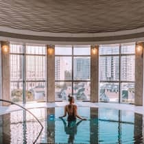 A woman swims in a circular spa pool in front of a wall of windows overlooking a city skyline