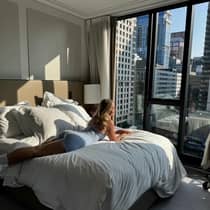 A woman lounges on a plush bed by a large window overlooking a city view
