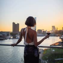 Person with bob haircut wearing a white halter-neck top and dark skirt stands silhouetted at sunset as they hold a glass of wine and overlook a city harbour