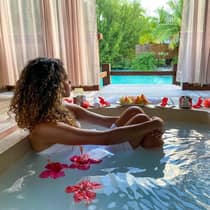 Person relaxing in a private spa bath with hibiscus flowers, overlooking a pool and tropical garden