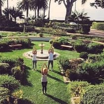 Three people stand with arms above head in yoga pose by fountain in green courtyard garden 