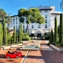 Young child rides in small red toy car in hotel courtyard with fountains, landscaped trees