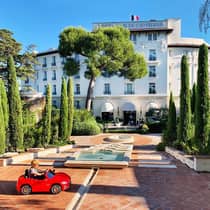 Young child rides in small red toy car in hotel courtyard with fountains, landscaped trees