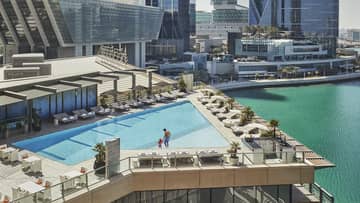 A wide shot of a man and child walking around a triangular pool overlooking the water and Abu Dhabi skyscrapers in the background.