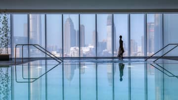 Silhouette of woman walking by blue indoor swimming pool, floor-to-ceiling window