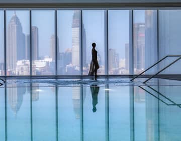 Silhouette of woman walking by blue indoor swimming pool, floor-to-ceiling window