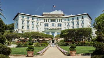 Two people walk down sunny stone path under large Grand-Hotel du Cap-Ferrat