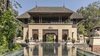 Person walking along the edge of a large reflective pool in front of a grand villa with traditional architecture surrounded by trees and outdoor seating