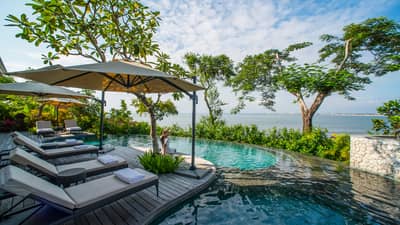 Lounge chairs with umbrella on wooden deck overlooking infinity pool, trees and ocean