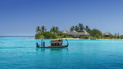 A wooden boat with passengers travels over clear turquoise water toward a tropical island spa with palm trees and thatched-roof villas