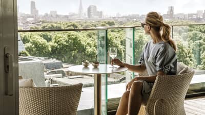 A guest takes in the city skyline while comfortably relaxing in a wicker chair and enjoying a drink on a balcony.
