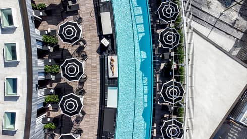 An aerial view looking down on a long, slightly curved pool and surrounding deck area, which includes tables with umbrellas, potted greenery and places where guests can recline next to the pool