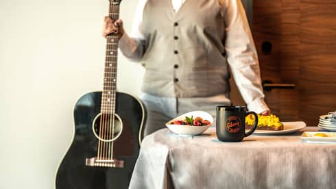 Person standing next to a black acoustic guitar, with a breakfast spread on a table featuring a "Gibson Garage" mug, fruit bowl, and scrambled eggs on toast.