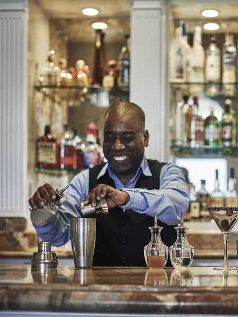 Smiling bartender pours liquor into martini cocktail shaker at bar