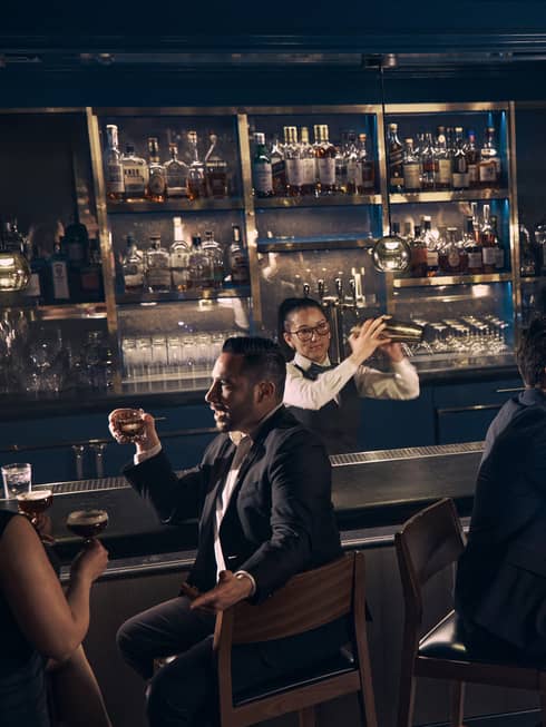 Guests chat over drinks at the bar at Prosperous Penny as a bartender shakes a cocktail shaker behind the bar.