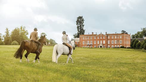 Couple rides horses across green English field to Four Seasons Hotel Hampshire building