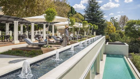 Main lounges on patio chair, woman stands by long fountain and ledge over plunge pool