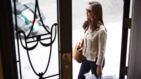 Woman wearing sunglasses, carrying designer bags walks along sidewalk past doors