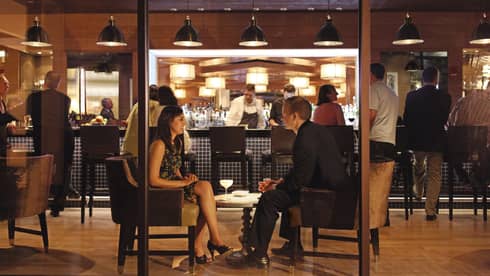 Beyond a glass wall sits a man in a black suit and a woman in a knee length black dress. They are having cocktails and chatting while the sleek bar bustles behind them. 