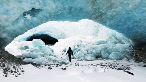Silhouette of person in snowsuit walking up to large chunks of ice, snow under large ice cap glacier