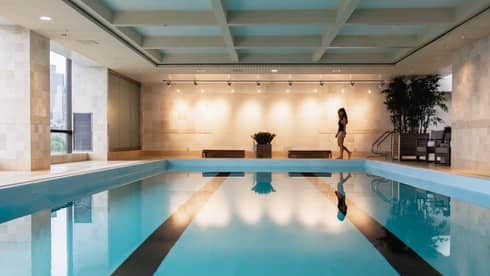 A woman walks in front of an indoor pool with a city view
