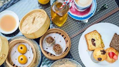 Breakfast table set with a variety of items: omelette and sausage, pastries in bamboo steamer baskets, drinks, etc.