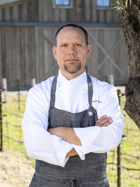A chef standing outside near a tree and in front of a barn.