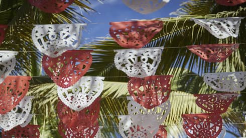 Looking up at rows of red and white Mexican Papel Picado banners hanging above, with palm trees and a sunny sky above them.