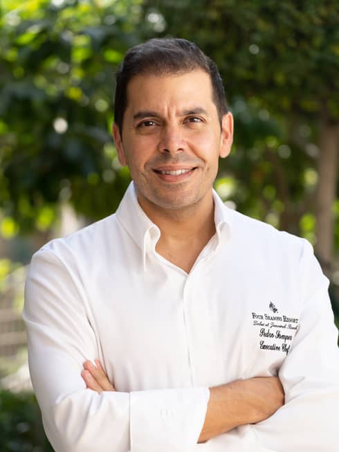 A chef in a Four Seasons uniform with his arms crossed in front of a canopy of foliage.