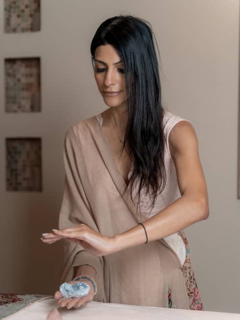 Woman with long dark hair wearing a tan-colored flowing dress hold a blue crystal in her hand while her other hand is suspended above it