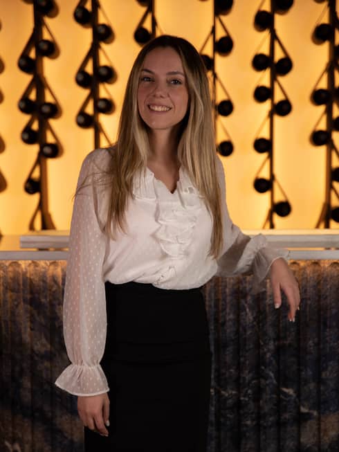 Monica Duarte in white ruffled blouse and black pencil skirt in front of gleaming restaurant wall