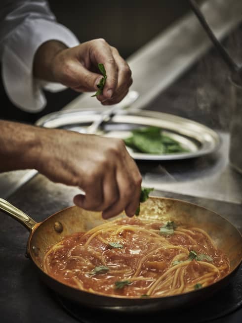 Fresh chopped basil is added to a steaming spaghetti dish in red sauce, prepared in a skillet on a stainless steel cooktop.